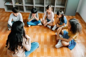 Elementary international students group sit cross legged in circle around the teacher and listening a story