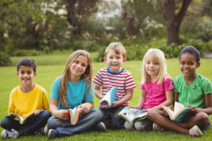 Smiling elementary school classmates sitting in grass and holding books